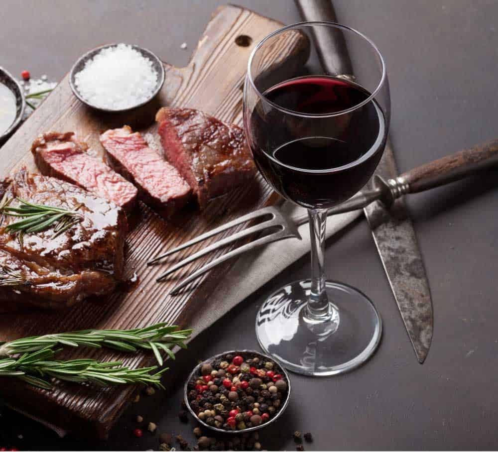 red wine with cooked steak on cutting board with herbs and spices
